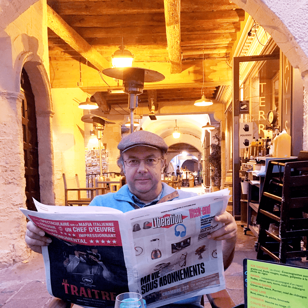 [a man in a hat reading a newspaper at an outdoor restaurant]