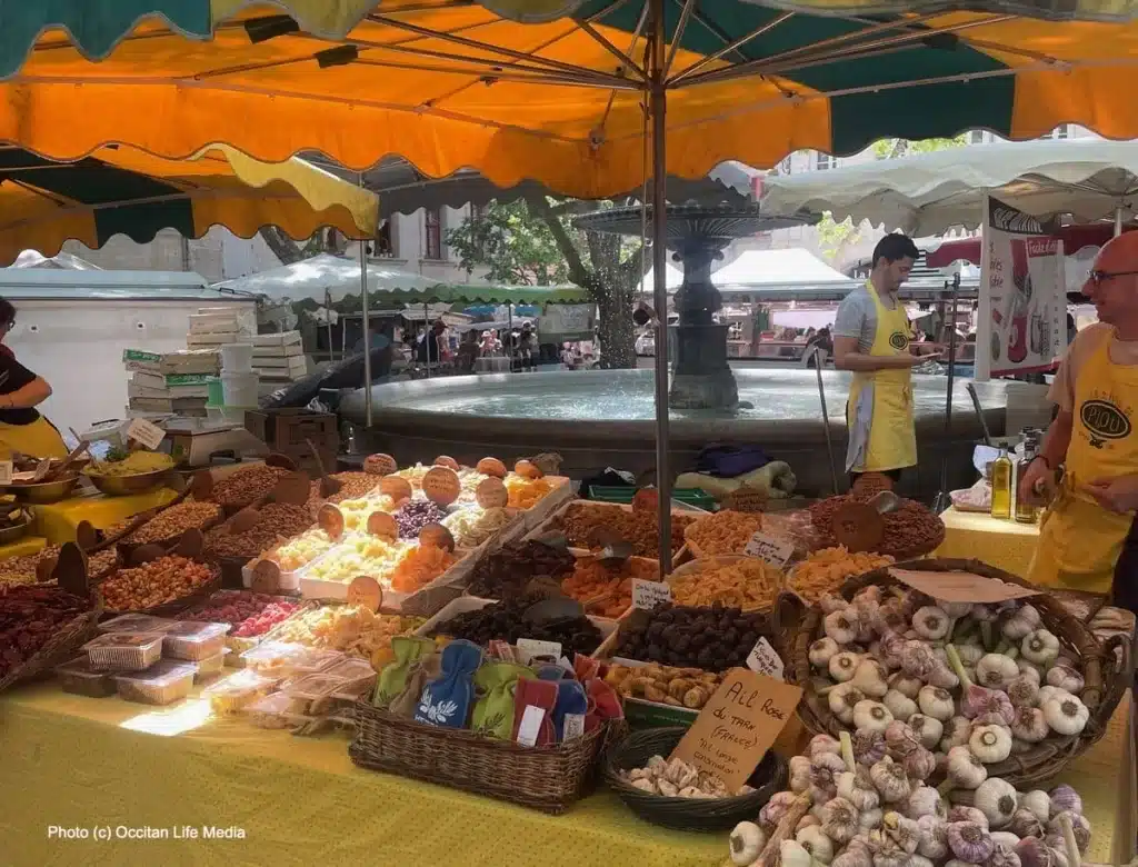 [an orange umbrella shades a display of vegetables with a fountain in background in Uzes France]