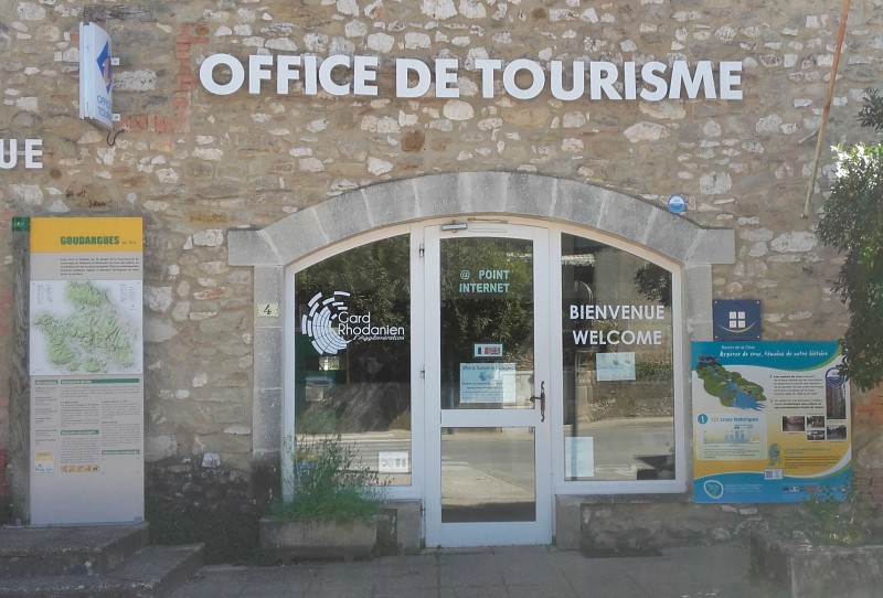 A photo of the outside of the tourism Office in Goudargues, in a stone made building with an arched glass door and some greenery. White letters over the doorway read: Office de Tourisme