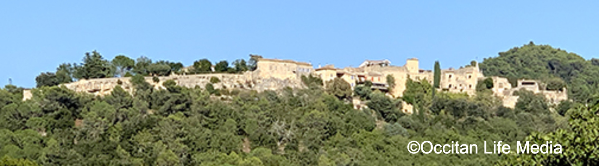 Up on a hillside, over trees, lies a village of cream-colored stone buildings against a blue sky.