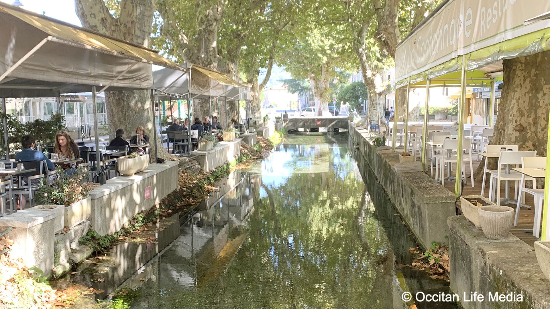 Canopied restaurants line a tree shaded canal in Goudarges in southern France, one of the best small villages in France People are seated and dining outside.