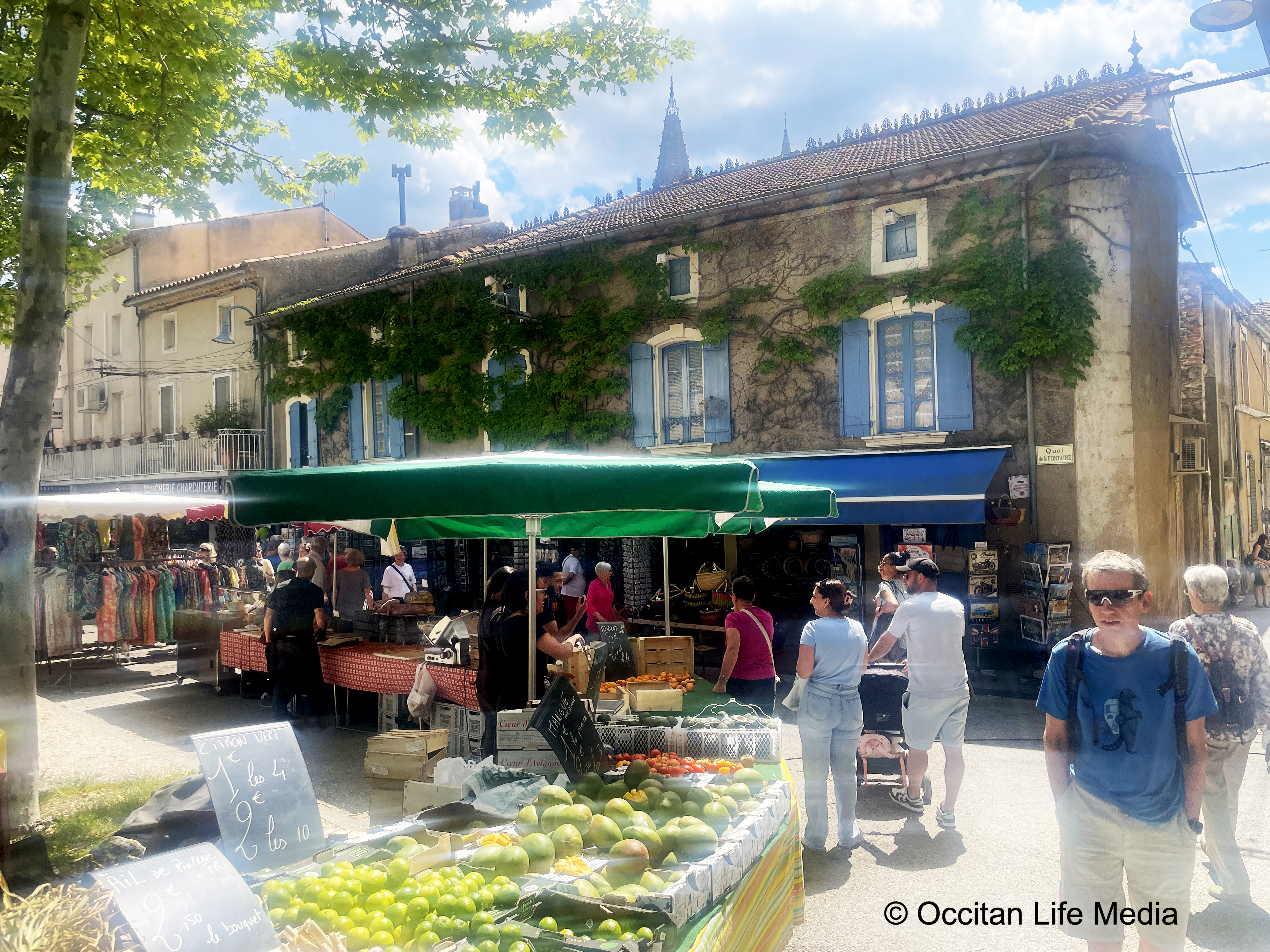 People stroll around a food market or marché in Goudarges France with sunny weather or wetter. Stalls display vegetables and fruits with colorful canopies. A building with blue shutter stands behind and a church spire can be seen.