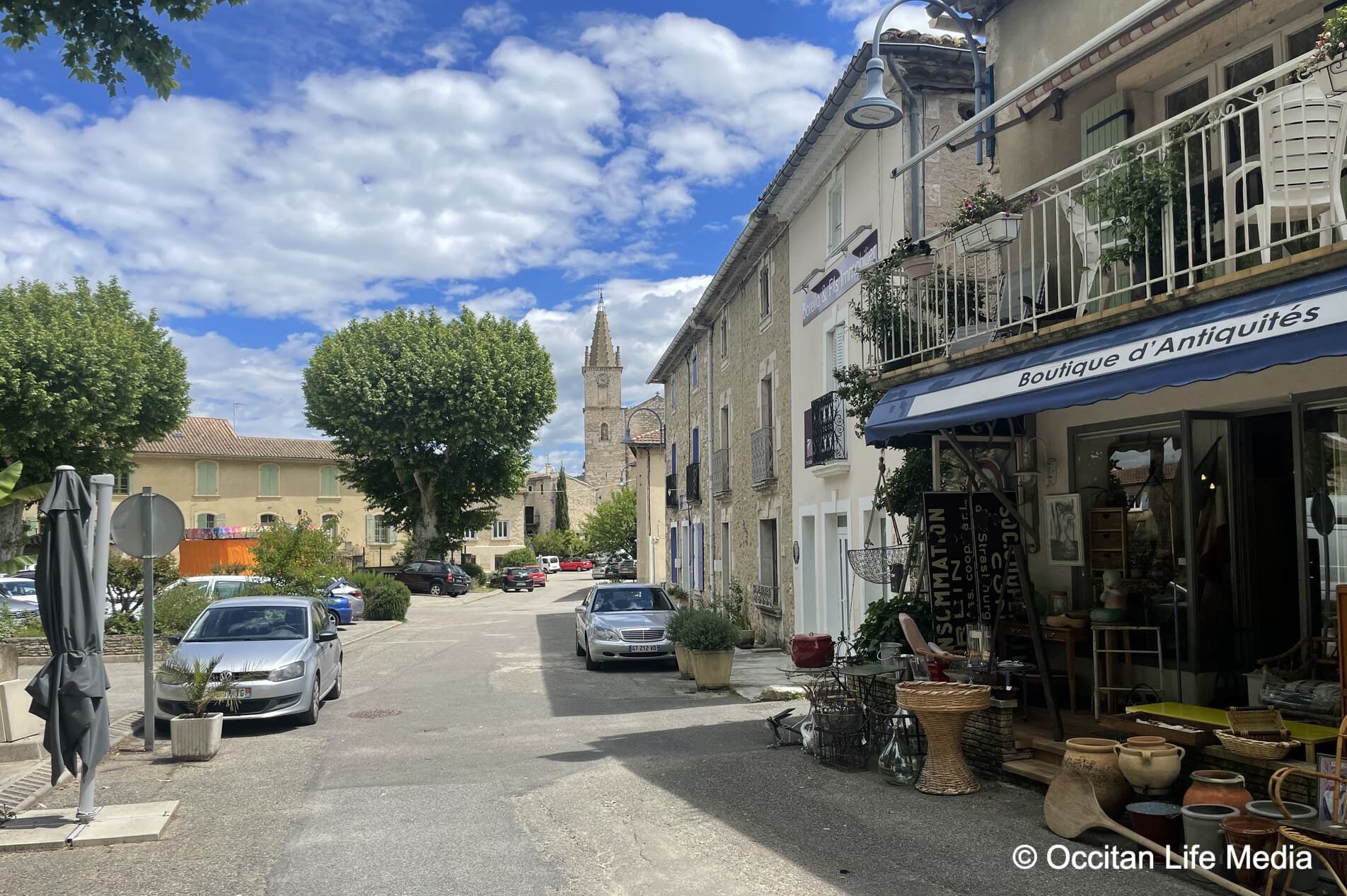 A view from the street of a charming village towards a church with a bell tower and spire against blue sky with white fluffy clouds. On rights is an antique shop with a display of household items.