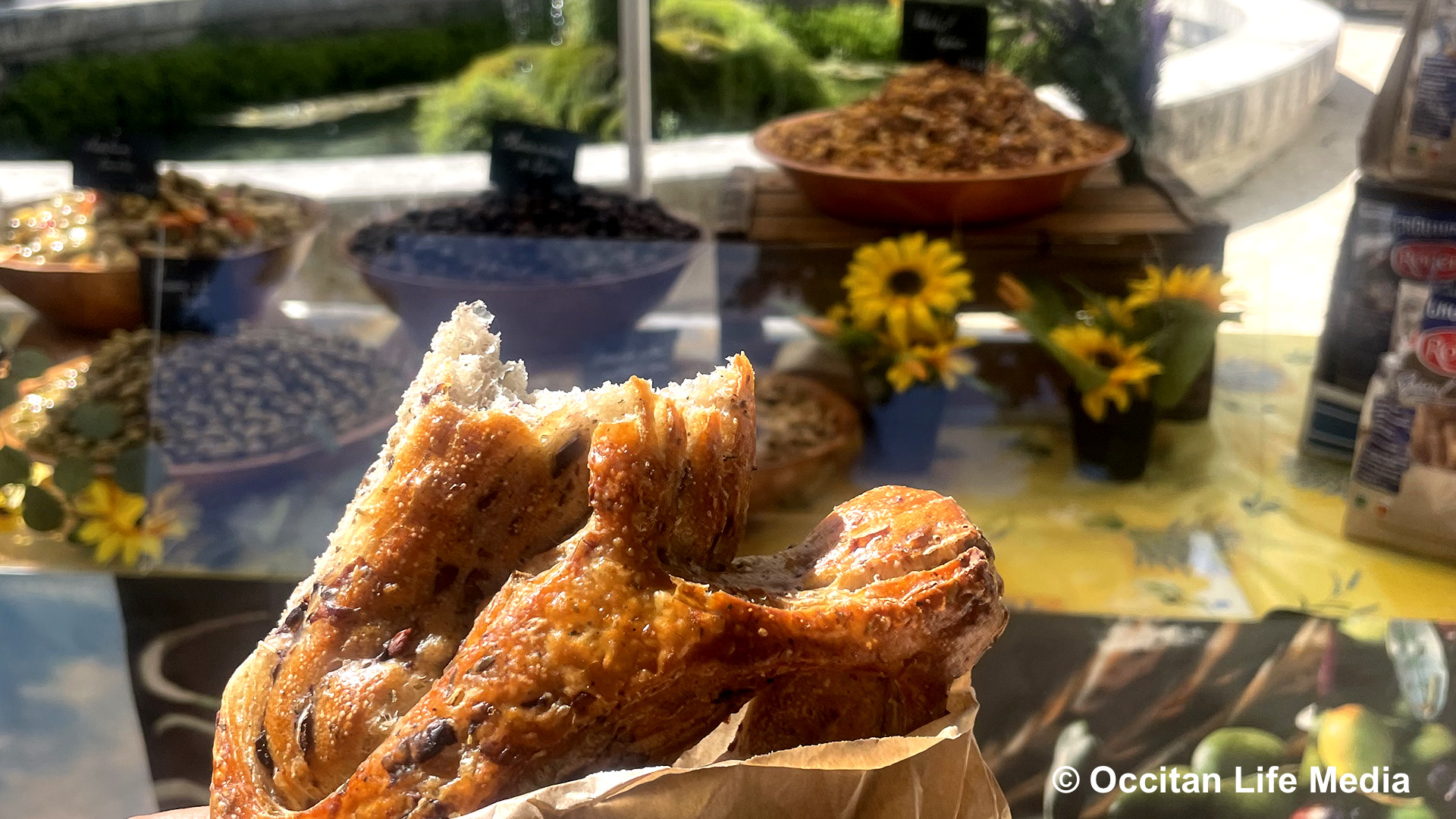 A partially eaten bread, called fougasse, which looks like a pretzel, int he foreground with a display of olives in bowls and sunflowers on a table in the background