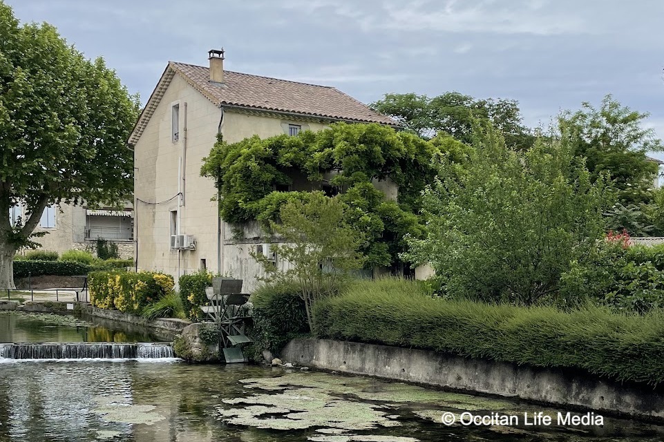 A canal flows past a cream colored building with red tile roof. Alongside the building is the remains of a watermill amidst greenery.